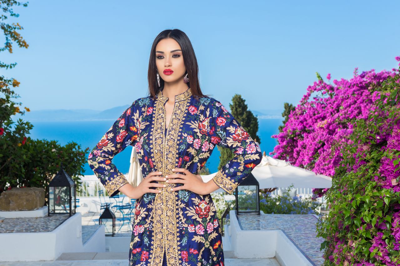 Woman in a floral shalwar kameez stands elegantly with colorful bougainvillea in the background.